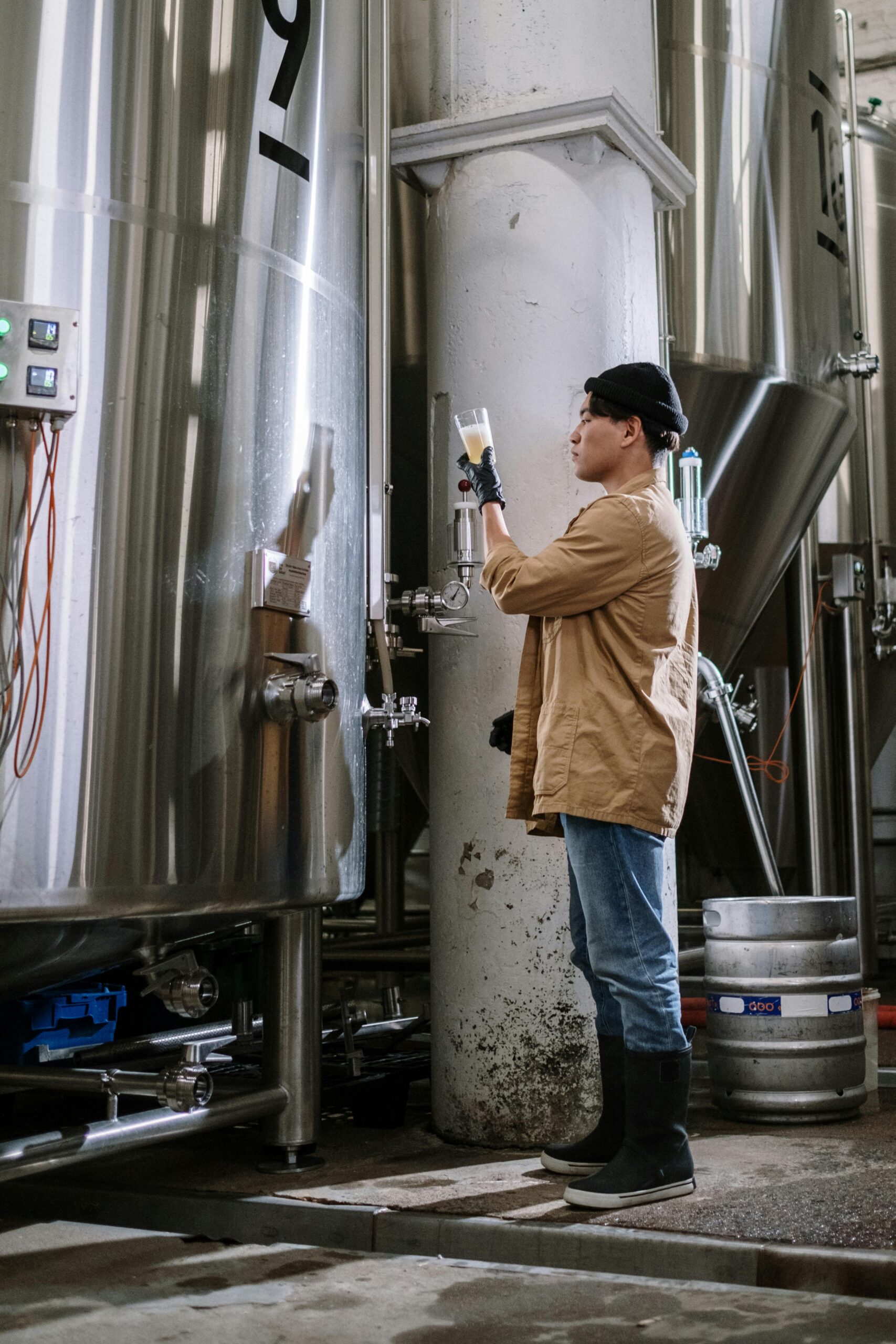 Asian brewmaster examining beer fermenter in a brewery. Industrial setting with stainless steel tanks.